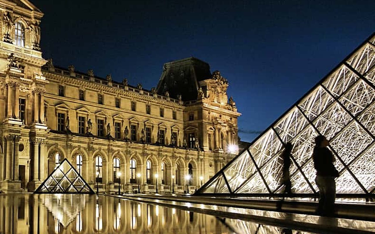 The glass pyramid lit from inside and the palace lit gold, both mirrored in the courtyard basin at night