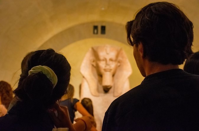 Visitors, seen from behind, in front of a granite sphinx in a lit crypt